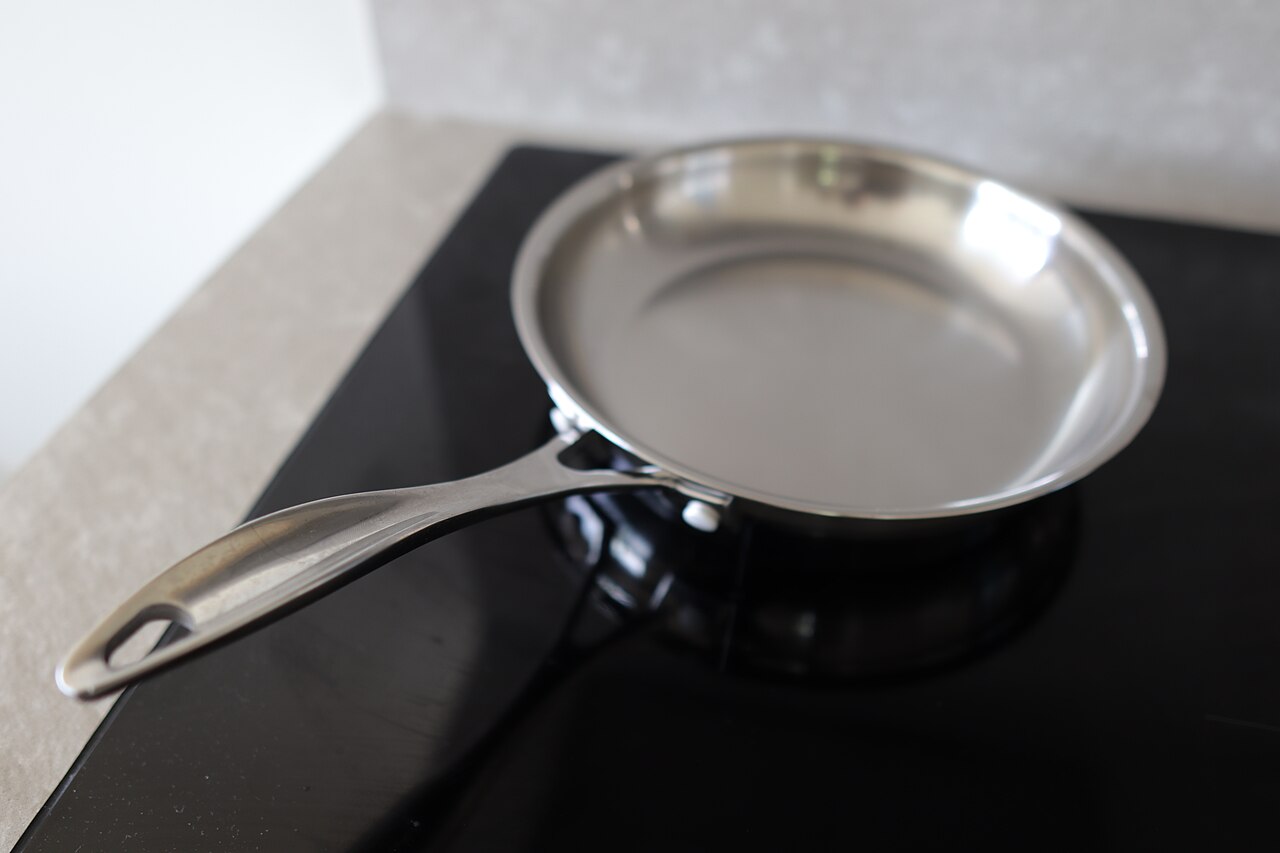 A silver-colored frying pan with a long metal handle on a black glass cooktop.